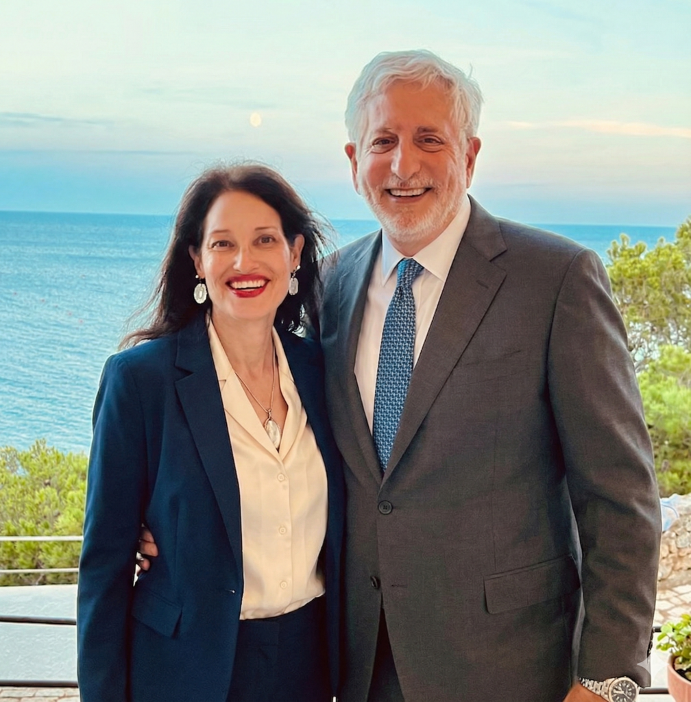 Portrait of Allan and Amy Baron, both in professional attire with Allan in a suit and tie and Amy in a navy blazer, posing on a balcony with a sea view